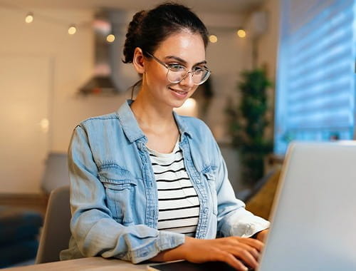 young woman student wearing glasses smiling typing on a laptop keyboard home office study