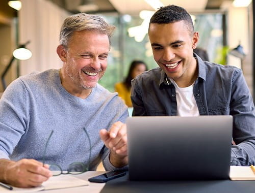 two men manager employee colleagues smiling looking at a laptop on a desk in an open office