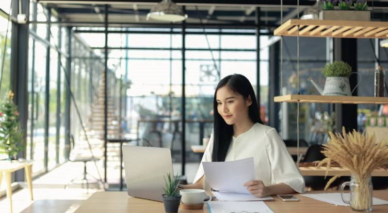 Young woman looking at paperwork in front of laptop in glass office